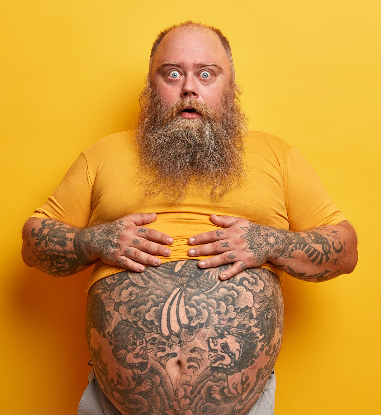 Shocked amazed hipster man keeps hands on belly with tattoo sticking out from t shirt, surprised to find out his weight, has long thick beard, poses against yellow background. Guy shows big abdomen
